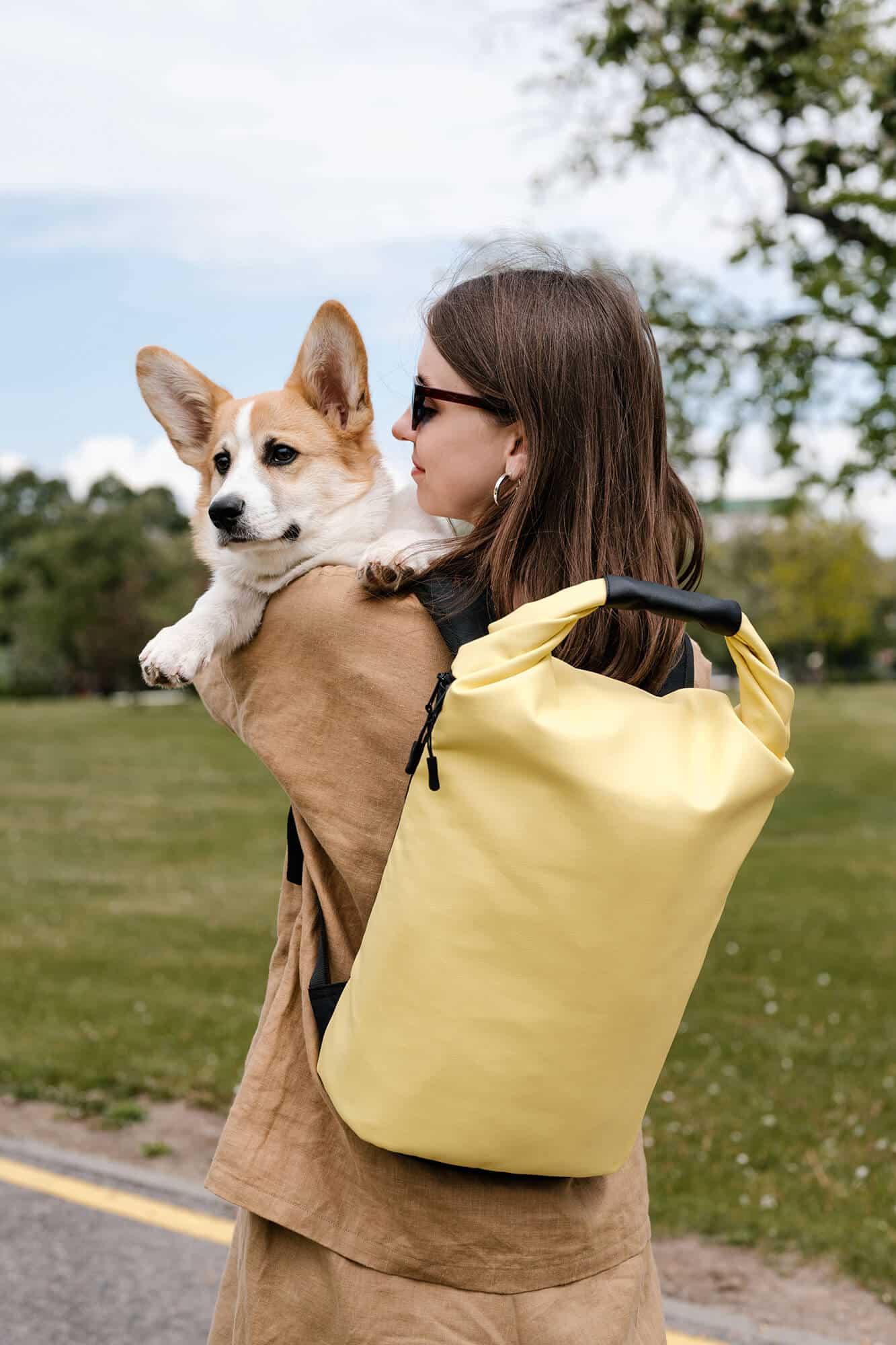A woman wearing a yellow backpack holding a corgi in her arms